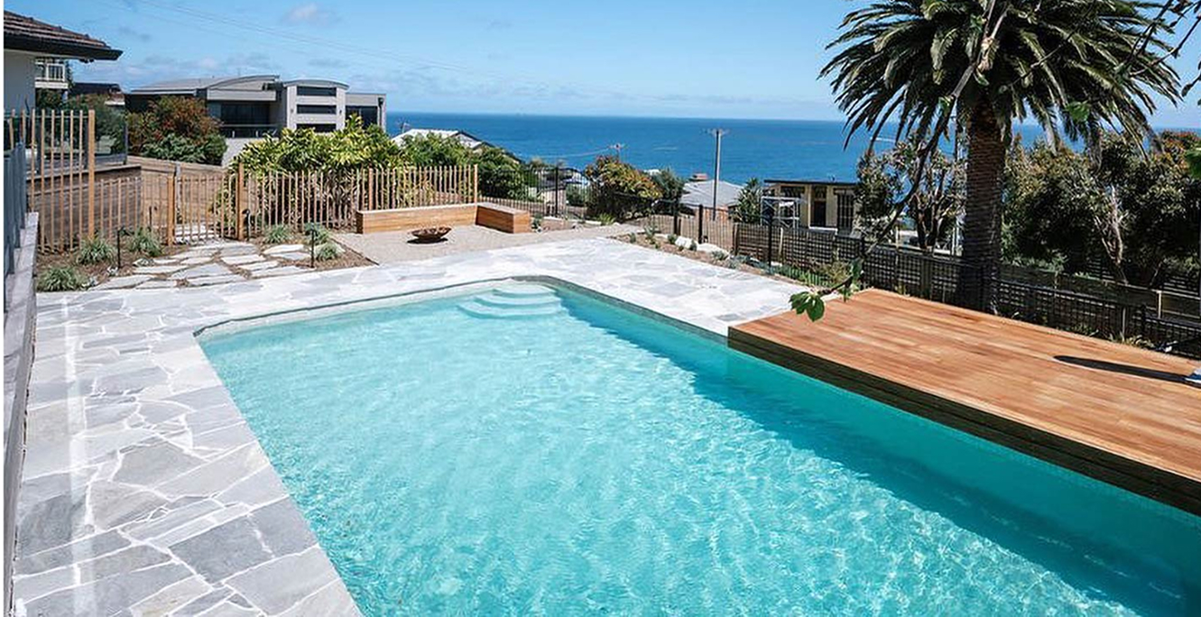 Sunlit coastal swimming pool with pale natural stone paving and ocean views